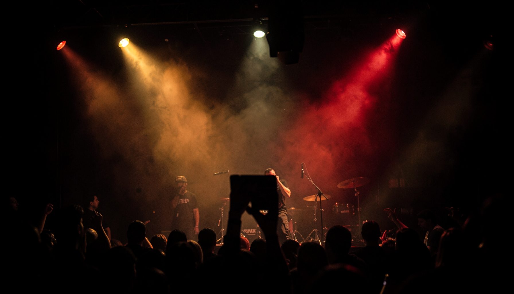 Dark atmospheric stage with warm amber and red spotlights cutting through haze, evoking the energy of a live hip hop show